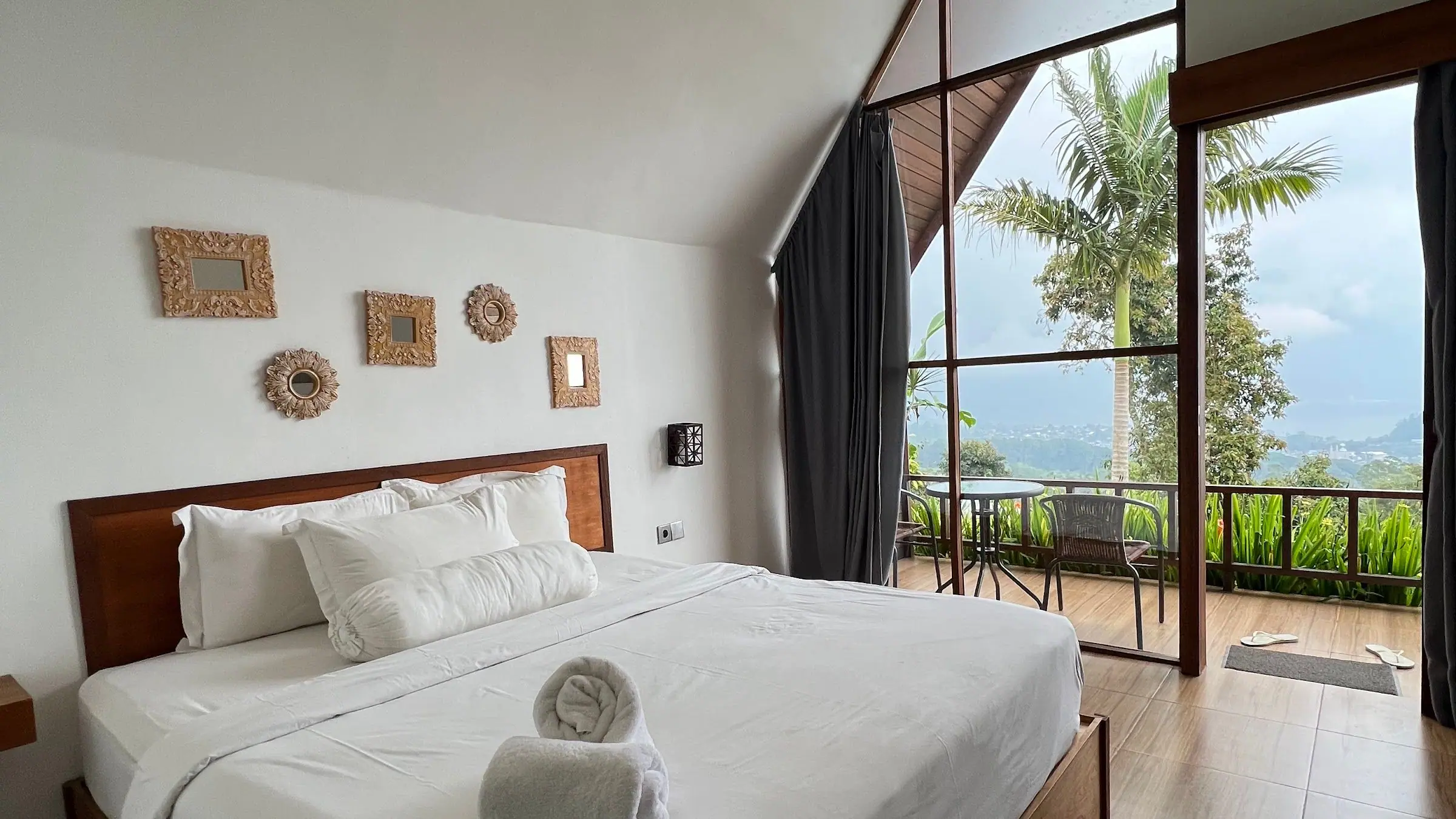 Inside an A-frame cabin at Catu Glamping: a made bed beside tall windows opening onto a balcony above the Bedugul hills and Lake Beratan